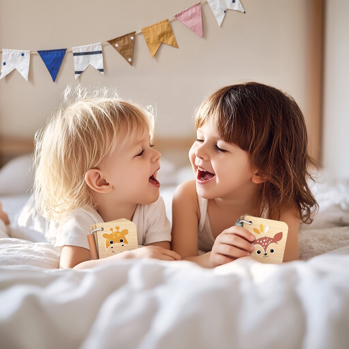 Two young children laughing together on a bed, each holding a Speedy Monkey Little Book — Jungle and Forest designs. Personalised wooden baby toys by NARA."