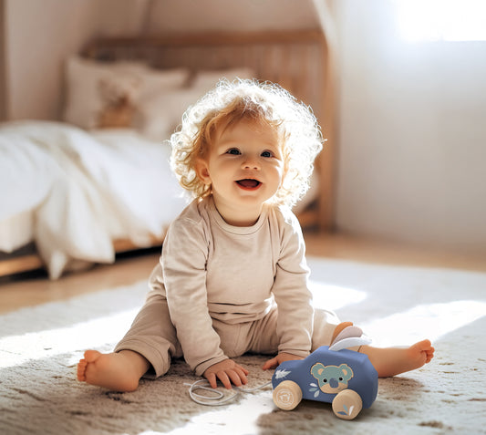 Baby with curly hair smiling and playing with a personalised Speedy Monkey Koala Pull Along Car on a nursery floor. Wooden baby toy by NARA.