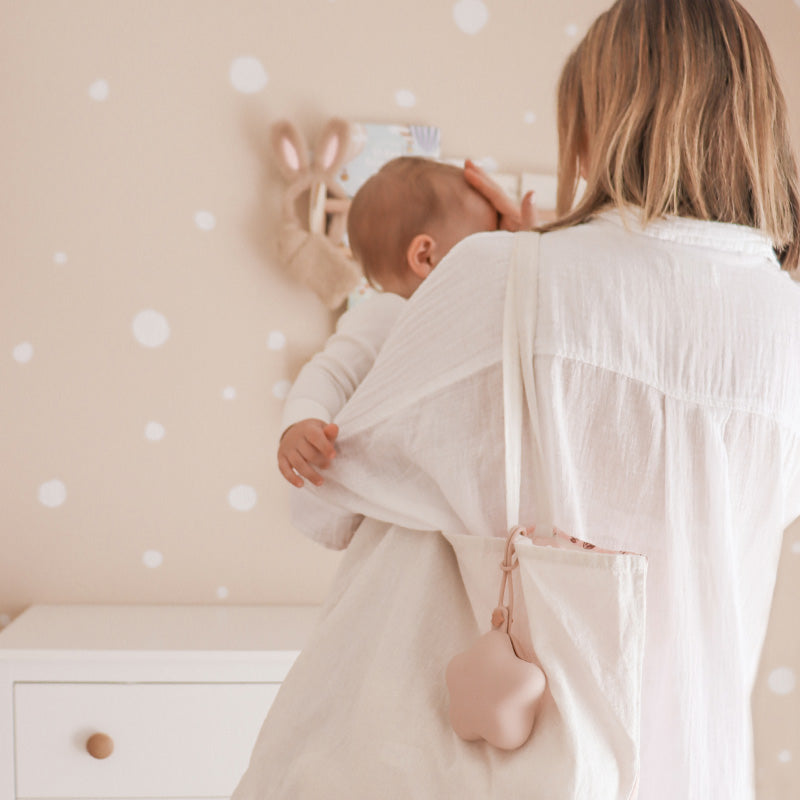 Mother holding newborn baby in a Scandi nursery — NARA personalised dummy clip visible on mum's bag