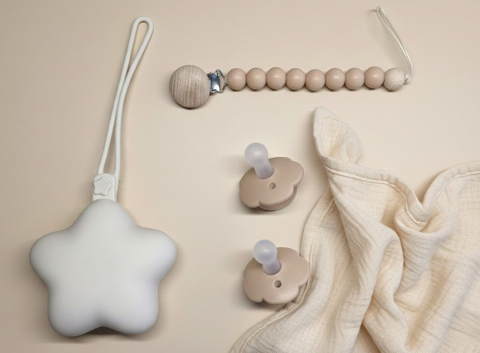 NARA Personalised Pacifier, Clip & Case Set flatlay — star-shaped silicone case, wooden bead dummy clip and two one-piece silicone pacifiers in Neutral Beige, styled on a warm beige background with a muslin cloth