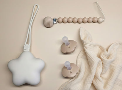 NARA Personalised Pacifier, Clip & Case Set flatlay — star-shaped silicone case, wooden bead dummy clip and two one-piece silicone pacifiers in Neutral Beige, styled on a warm beige background with a muslin cloth