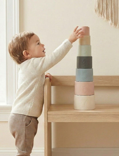 Toddler stacking NARA silicone stacking cups into a tower on a wooden table — set of 7 numbered cups in muted Scandi tones