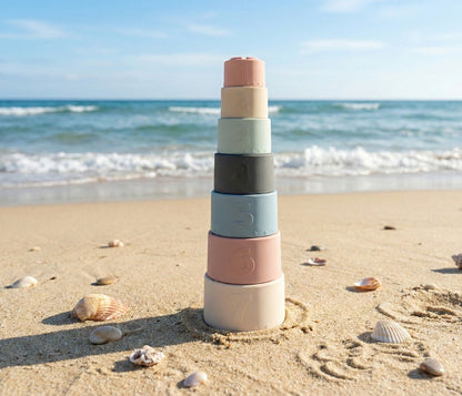 NARA silicone stacking cups stacked as a tower on a sandy beach with seashells — 7 numbered cups in muted pastel tones